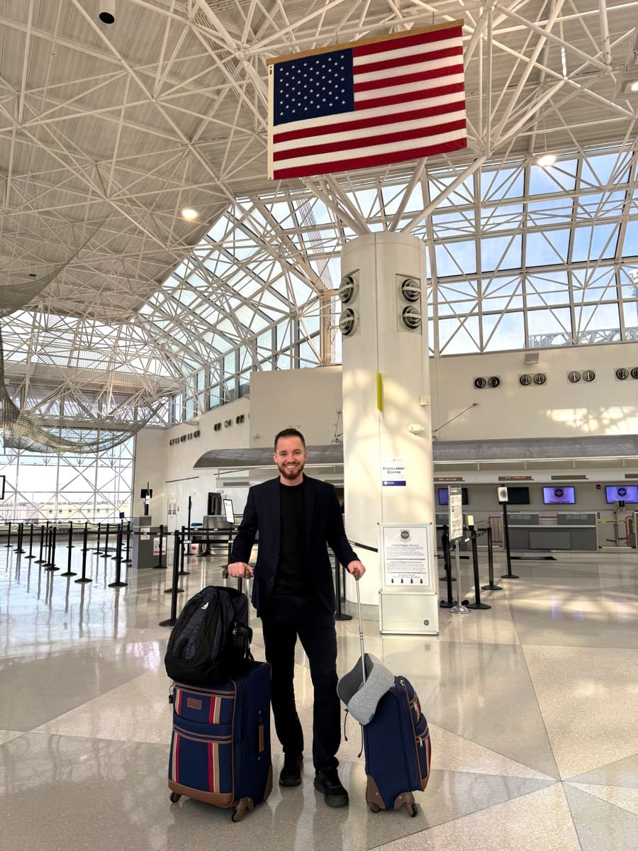 Bruno Dornelles, fundador da Immigrow, com as malas em um aeroporto nos Estados Unidos, sob a bandeira americana